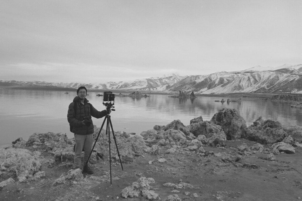 Black and white photo of Beihua Guo standing beside a camera on a tripod on a lakeside with mountains in the background.