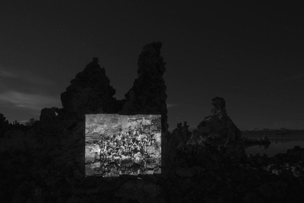 A black and white photo of a rock formation by Mono Lake at night. A photo of people gathered for a rehydration ceremony is projected on the rock formation.