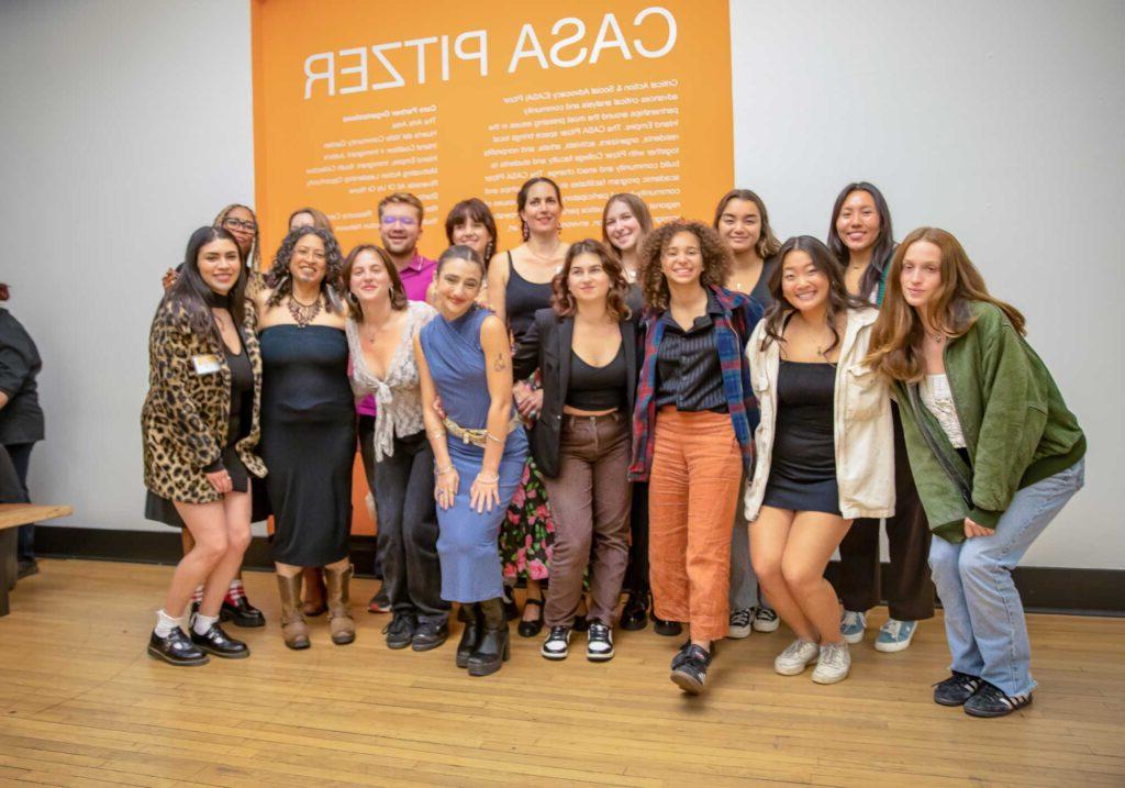 CASA Pitzer students and staff take a group photo in front of an orange wall with CASA Pitzer's name and mission in white text.